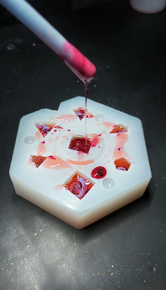 You see a white dice mold shaped like a hexagon on a dark background. The mold is filled with resin in orange and red with purple swirls. A stiring stick covered in red resin is visible, a thin streak of resin is dripping from the stick into the mold.