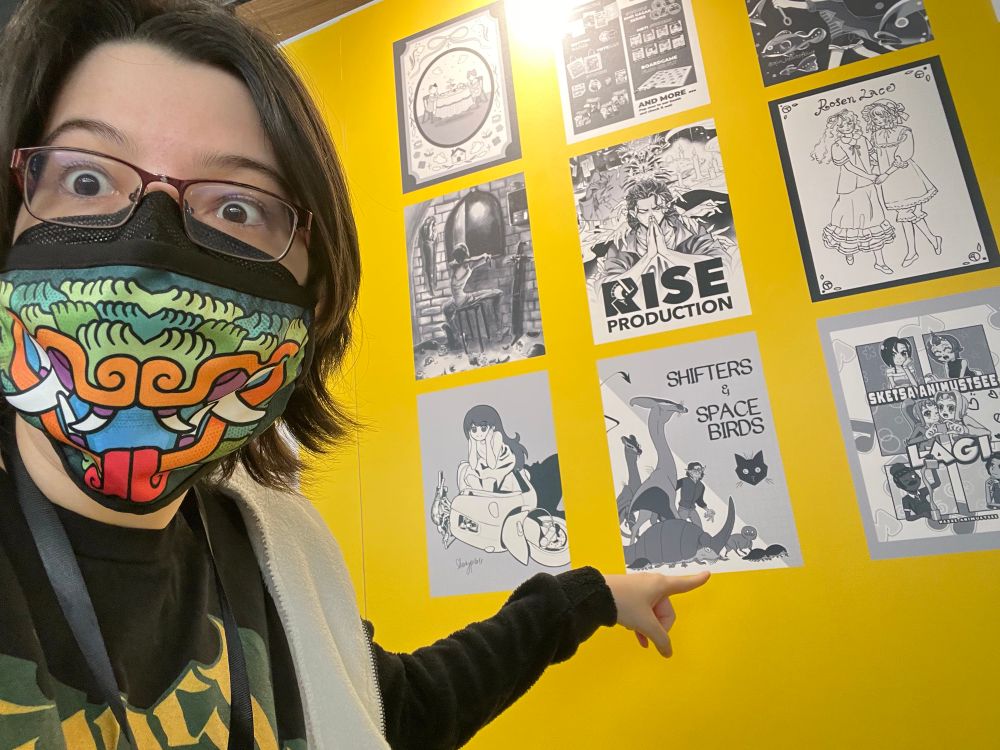 Person with rectangular glasses and a mask featuring a Maya style monster mouth pointing at a board with several posters on it, particularly one labelled “shifters and spacebirds”.