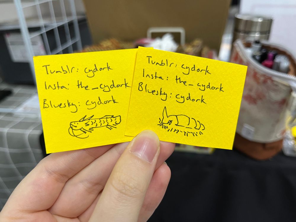 Yellow pieces of card with social media details written on them, and small silly drawings of a dragonfish and isopod-like alien. These were placeholders for business cards.