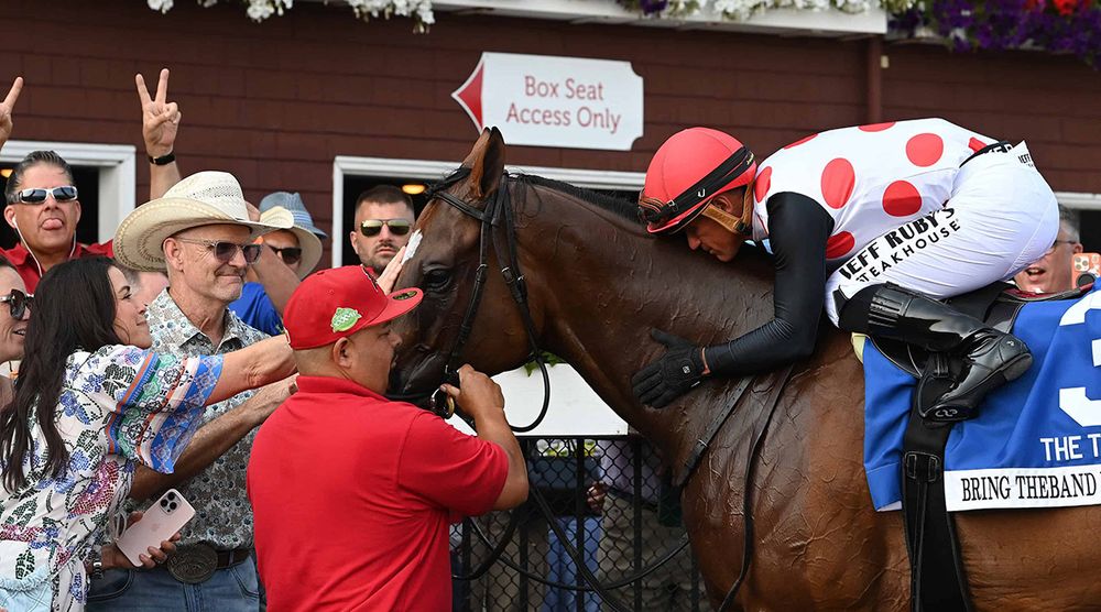 Bring Theband Home, bay Florida-bred racehorse, stands in the middle of the winner’s circle at Saratoga, showered with attention by his connections after winning the 2025 $300,000 Troy (Grade 2). On Bring Theband Home’s back, jockey Javier Castellano leans down, gently hugging the gelding after their well-earned victory. (Photo: ©Susie Raisher)