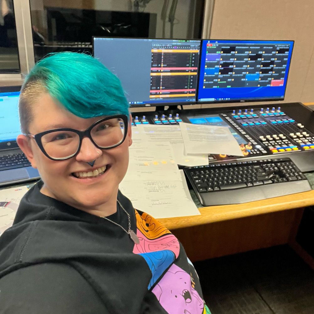 A non-binary person with short blue hair and an adventure time tshirt is sitting in a radio studio surrounded by notes, clocks, and programming language. The soundboard and monitor is behind them. 