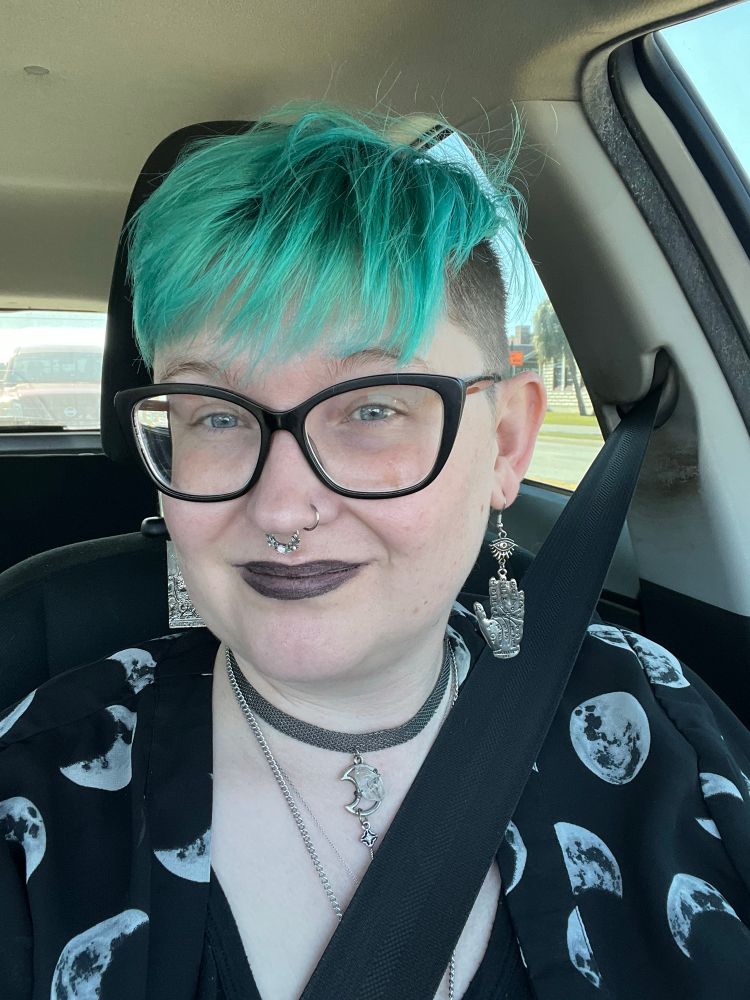 A pale-skinned nonbinary person with short greenish blue hair, black glasses, and black lipstick smiles into the camera. They’re wearing an over shirt with moons on it. 