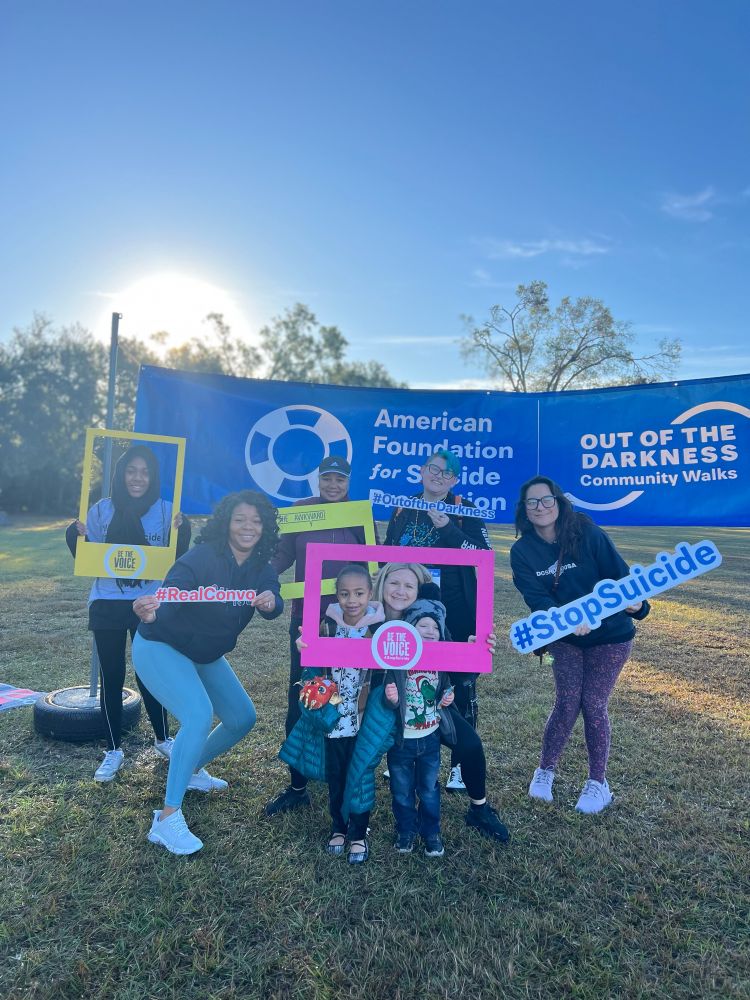 A group of six adults and two children decked out in warmer clothing for the cold morning pose in front of a long blue American Foundation For Suicide Prevention Banner for the Out of the Darkness Community Walk. They’re holding various signs that say things like stop suicide, real convo, and be the voice. It’s a crisp December day at the park and the sky is very blue, the sun just barely up over the horizon. 