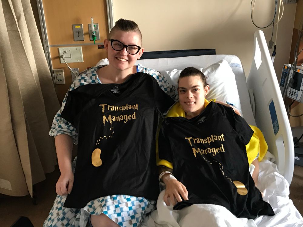 Two smiling siblings in hospital gowns on a hospital bed. They’re holding up matching shirts that have gold foil kidneys, a little trail of footsteps, and the words “transplant managed” in the Harry Potter font. 
