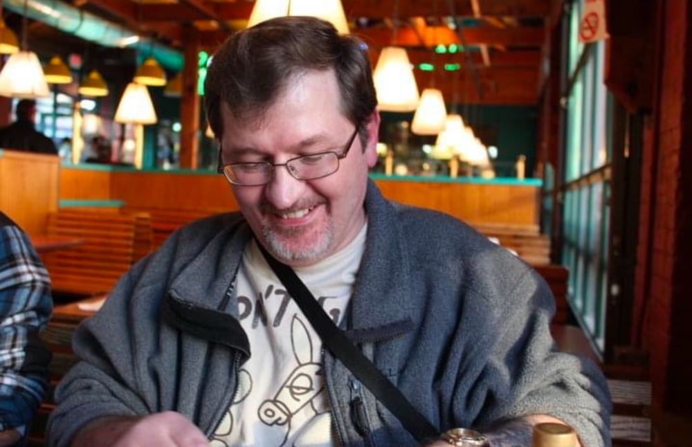 A middle aged white man with short brown hair and glasses wears a white tshirt and a gray zip up jacket and is sitting at a restaurant in wheeling, West Virginia. 