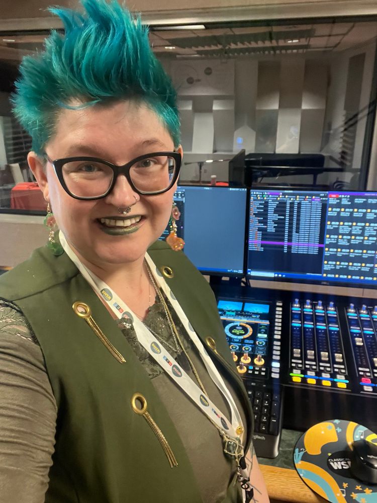 A person wearing an olive green and gold outfit with shirt blue hair and glasses stands in front of a brightly lit digital soundboard inside the WUSF studio during their fiscal year end pledge drive. (And hopes you donate!) 