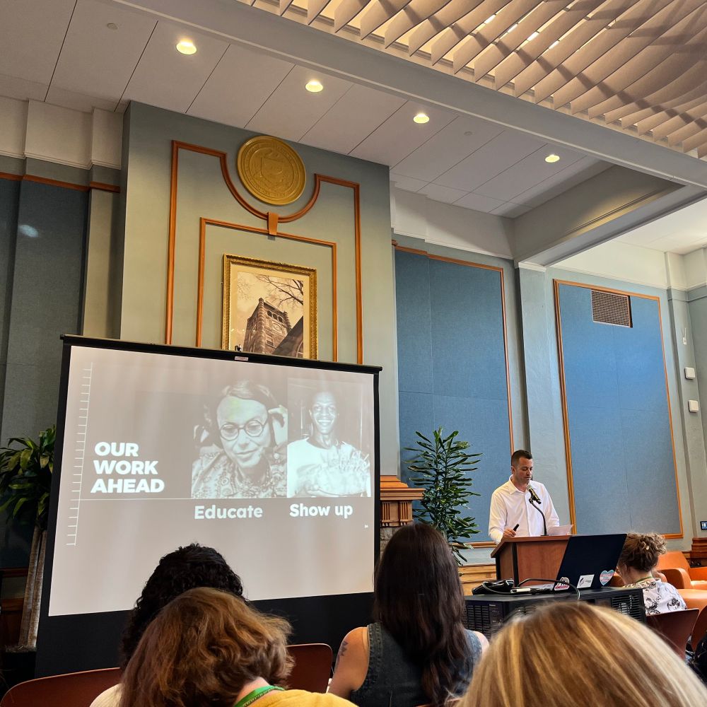 K.J. Rawson stands in front of a projector screen. It reads "Our Work Ahead: Educate, Show up" alongside photos from the Digital Transgender Archive. 