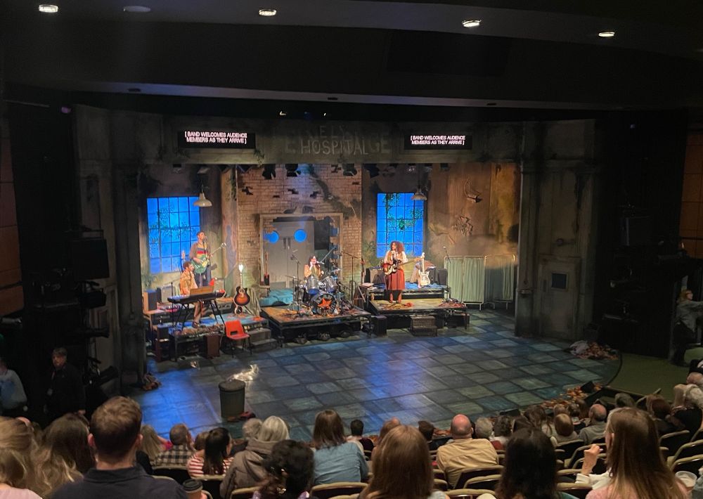 Stage of ‘Housemates’ at the Sherman Theatre in Cardiff. Some of the cast are playing instruments and singing before the play starts.