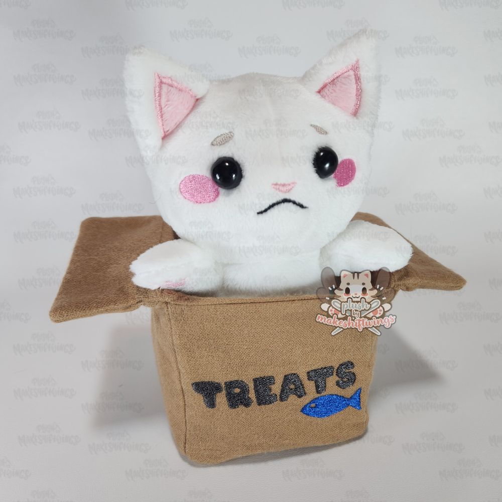 A plush of a white sad cat in a cardboard box labeled "treats"