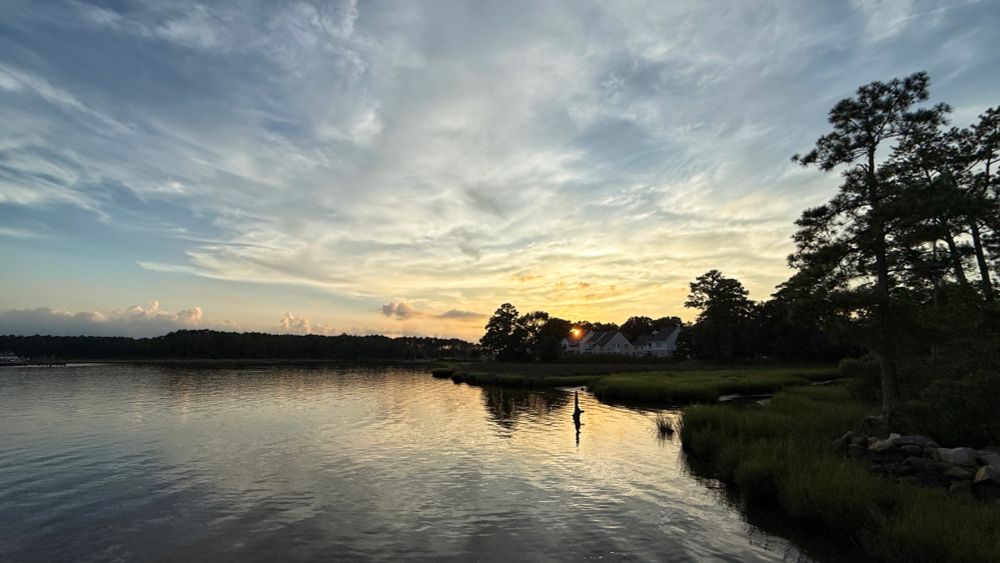 The setting sun shines through the distant trees near the horizon. The clouds take on the hues of the orange/yellow sunlight against a blue sky and are reflected in the river waters below. The far shoreline and trees on the right are silhouetted against the sky.