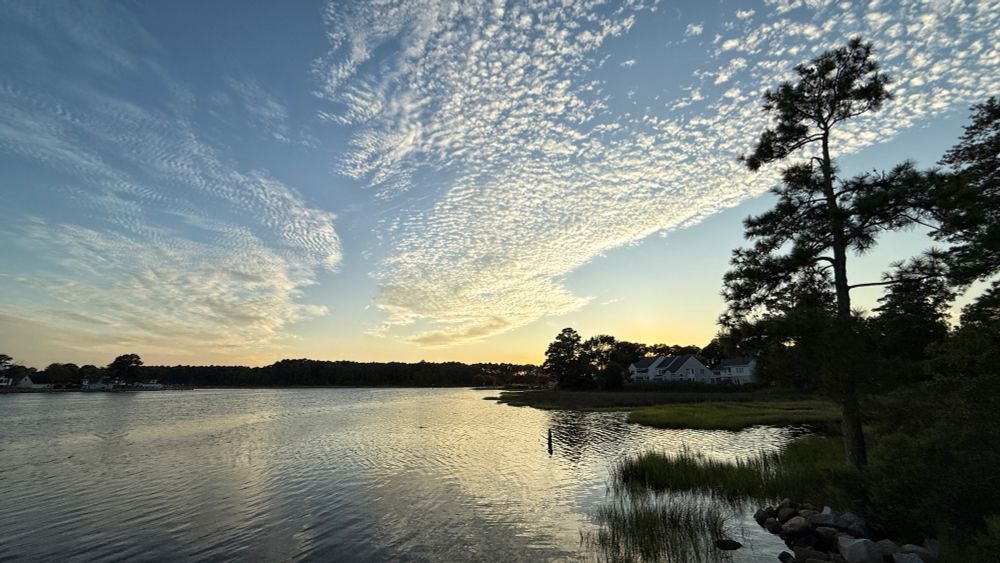 The setting sun illuminates the high scattered clouds, which are reflected in the river water below.