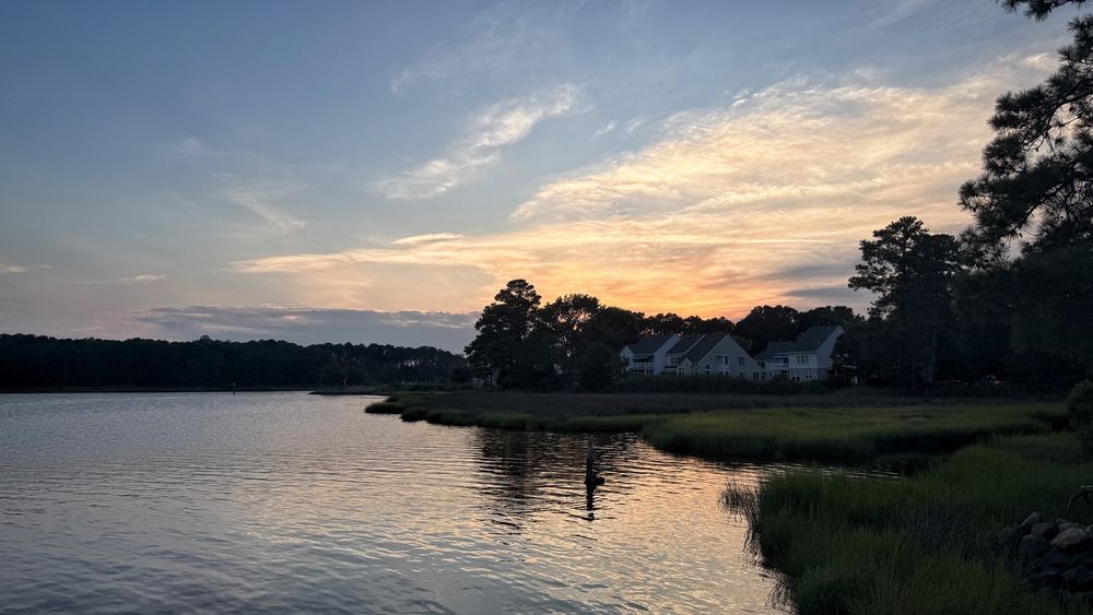 The setting sun casts a yellow and orange light on thin clouds. The river waters are calm and reflect the golden light.