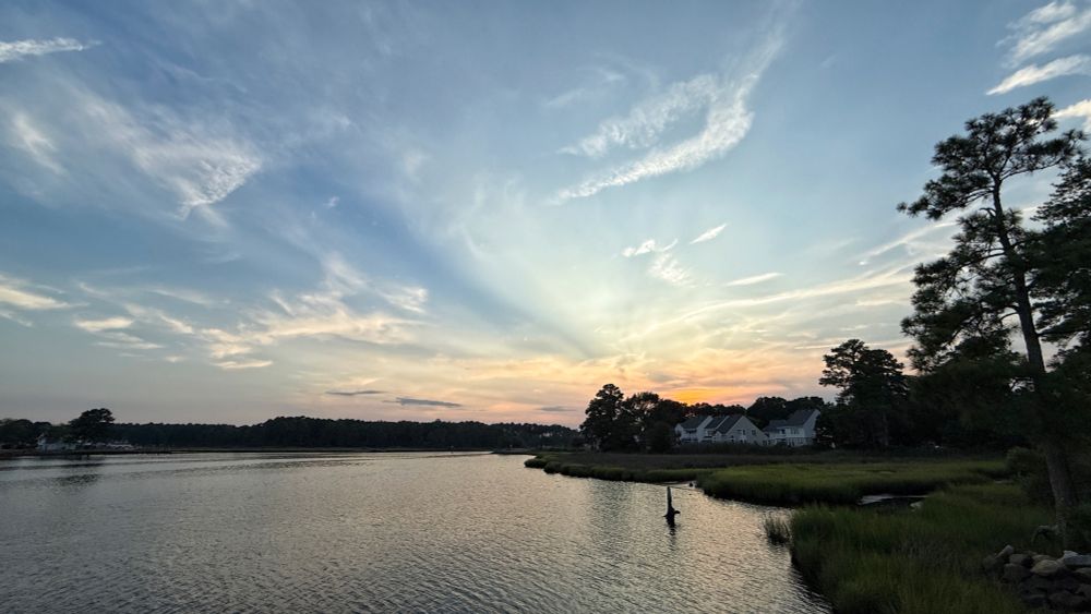 The sun has disappeared behind the distant trees. An orange glow colors the clouds near the horizon. High, white clouds sweep across the blue sky above. The far shoreline and trees on the right are silhouetted against the dusk sky.