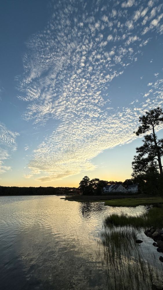 The setting sun illuminates the high scattered clouds, which are reflected in the river water below.