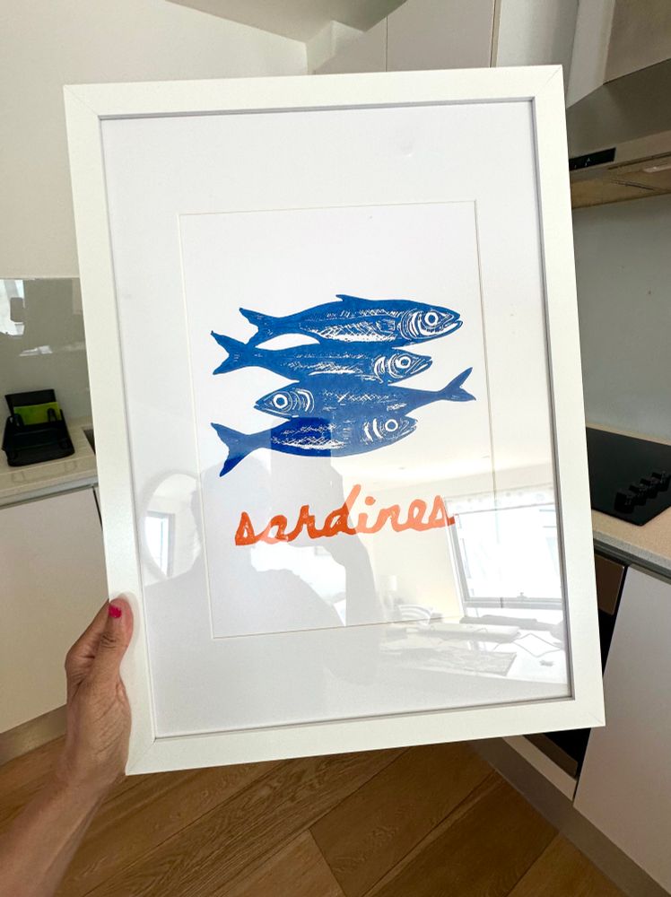 Holding up a framed handmade art Lino print of 4 sardines with ‘sardines’ in orange underneath them. 