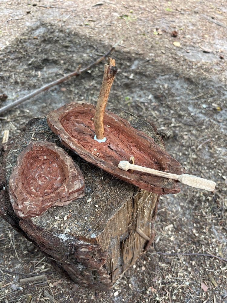 Handgeschnitztes Holzspielzeug auf einem Baumstumpf: Ein einfaches Boot aus rötlich-braunem Holz mit einem Mast aus einem Ast, befestigt mit Leim. Daneben liegt ein geschnitztes Ruder oder Paddel aus hellem Holz. Im Hintergrund ist Waldboden mit Spänen und Rinde zu sehen