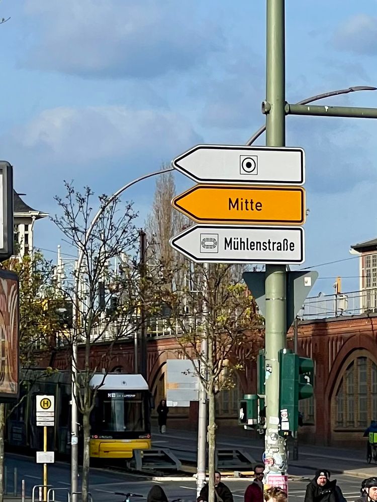 Ein Straßenschild mit drei Pfeilen nach links. Im oberen ist auf weißem Grund ein Kreis in einem Quadrat mit einem Punkt in der Mitte. 
Im mittleren steht auf gelbem Grund das Wort Mitte. 
Im unteren auf weisem Grund ein Piktogramm für Arena und das Wort Mühlenstraße. 