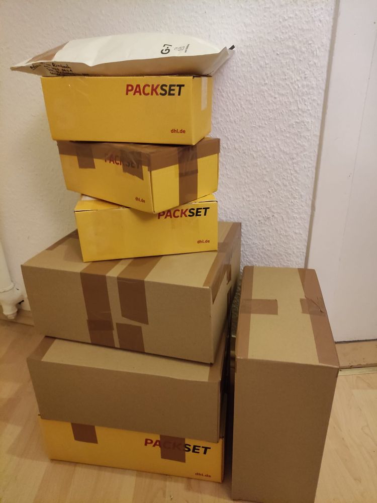 Foto von einem Stapel gepackter und zugeklebter Kartons, die auf dem Fußboden stehen.
Man sieht insgesamt sieben Pakete und einen großen Versandumschlag, der oben auf dem Stapel liegt.