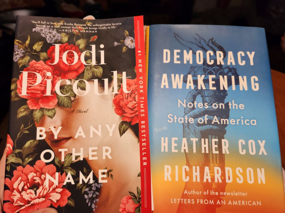 2 books. Just finished, and on the left is "By Any Other Name" by Jodi Picoult. Just started and on the right is "Democracy Awakening" by Heather Cox Richardson.