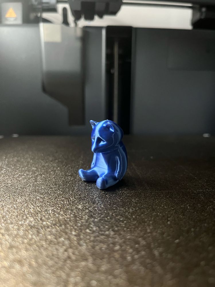 Small, blue, 3D printed raccoon sitting on the printer's build plate, slouched with a sad expression