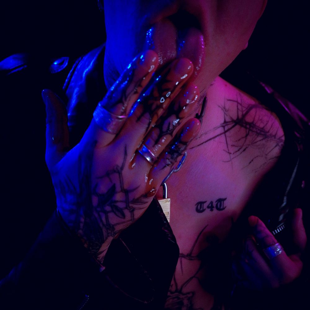 A dark colour shot of Lex, a white tattooed masculine person with a shaved head, licking Huberd's shoe grease off His hand. He's shirtless and wearing a black leather jacket.