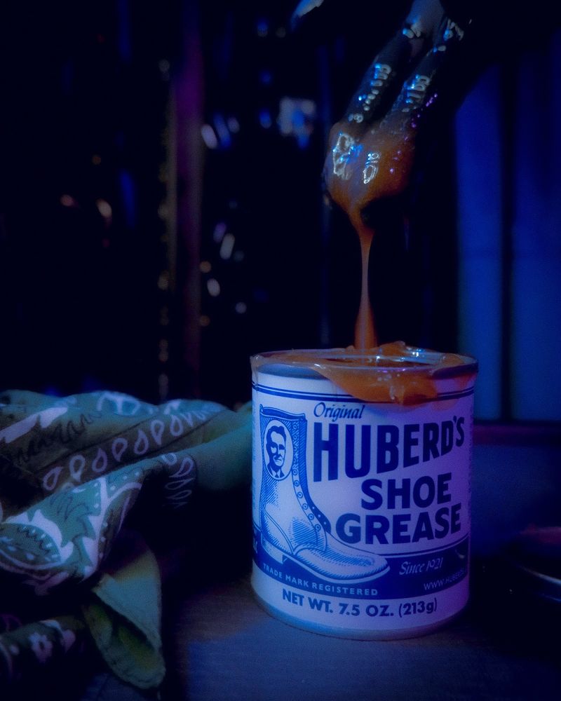 A soft colour shot of a can of Huberd's shoe grease next to a hunter green hankie. There is a nitrile gloved hand above the can dripping melted shoe grease into it.