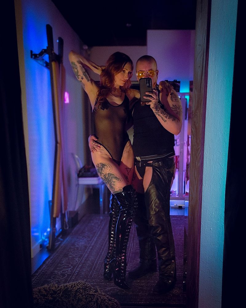 Dominus, a tall white tattooed masculine person with a shaved head, and Goddess, a tall white tattooed person with long red hair, take a mirror selfie in a dungeon. Dominus is wearing black leather chaps, a black tank and a black leather shoulder harness. Goddess is wearing a black mesh bodysuit and black thigh high vinyl boots.