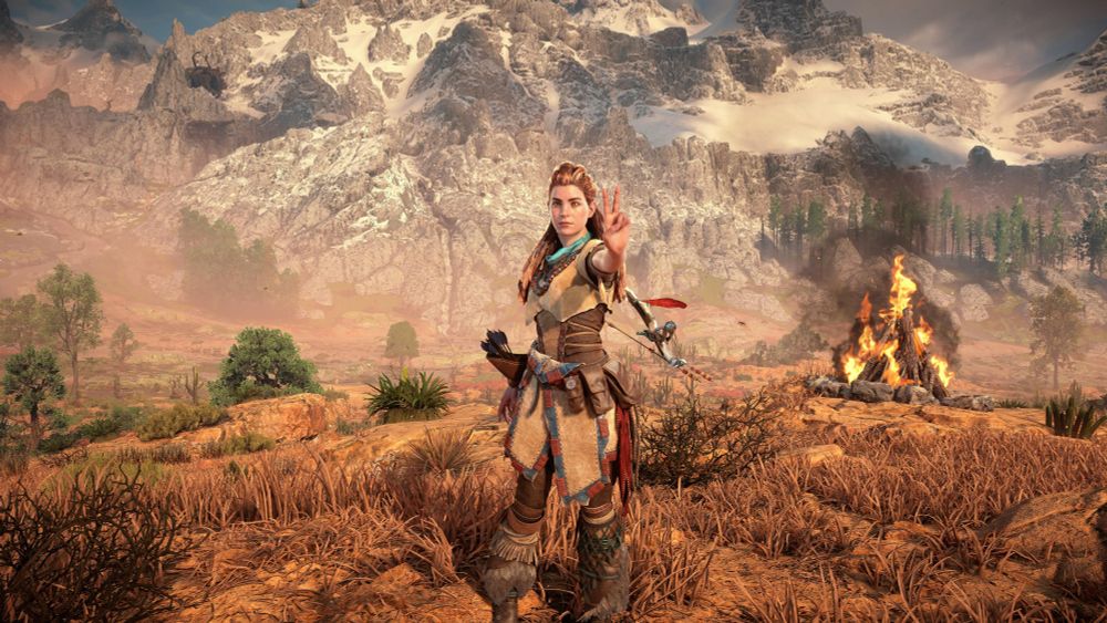 Dieser Screenshot zeigt eine Szene aus dem Videospiel Horizon Zero Dawn. Es ist eine karge braune Wüstenlandschaft mit einigen wenigen Pflanzen zu sehen, aus der sich im Hintergrund mächtige verschneite Gebirge erheben. Im Vordergrund steht mittig im Bild die rothaarige Protagonistin Aloy, die den linken Arm nach vorne streckt und mit zwei Fingern das Victoryzeichen macht. Links hinter ihr sieht man ein sehr grosses Lagerfeuer. 