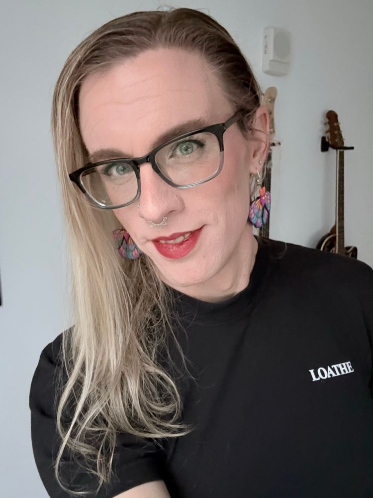 Woman with blonde hair and blue eyes in a black tshirt with the word “loathe” in white capital letters on the chest standing in front of a white wall. A few guitars are barely visible hanging behind her. 