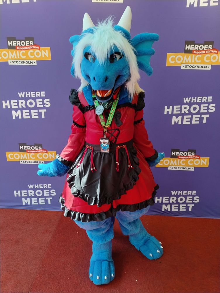 A blue dragon fursuiter standing by a photo background of Comic Con Summer Edition in Stockholm wearing a red and black dress, with a red collar with a grey tag reading "Roxee" and the pronouns "she/her". The fursuit has earfins, white horns, and icy blue hair fading to orange, and white claws on digitgrade feet paws. The underside is a burgundy carpet.