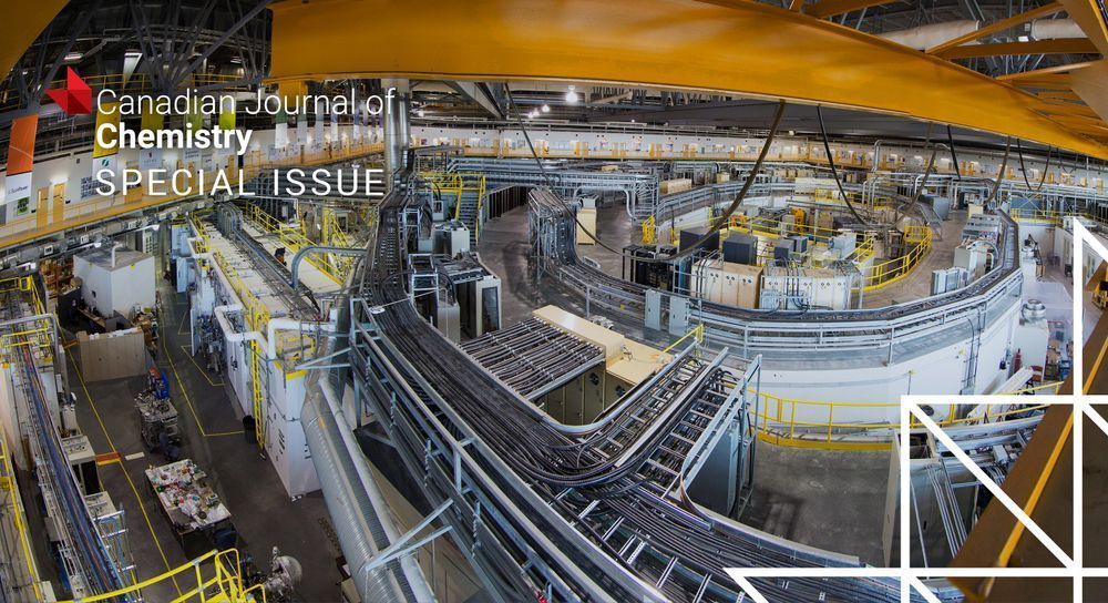 A large circular particle accelerator facility with complex metal piping and yellow safety railings inside an industrial building.