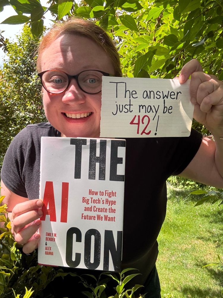 Me holding up the book “The AI Con”, with a little piece of paper matching in red and black colors with the book, “The answer just may be 42!”