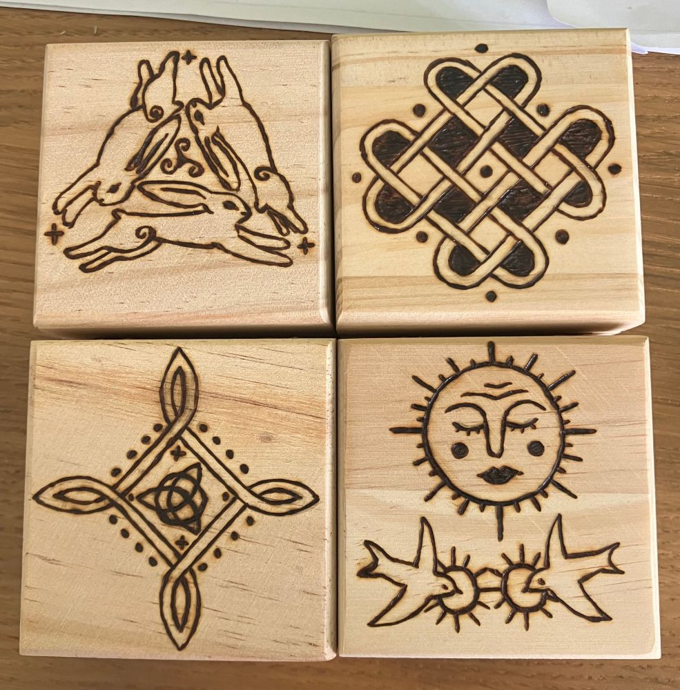 the tops of four square boxes each with various celtic motifs wood burnt on them. the top left is three hares in a triangle, ears touching as they run with a triskele between them. the top right and bottom left are two celtic knots. the bottom right is a sun with closed eyes and pouty lips above two swallows flying with a small halo around their heads. 