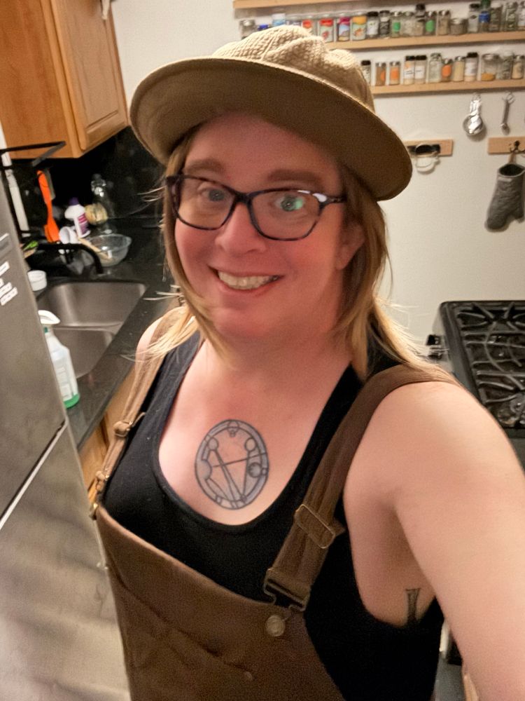 me, lookin' really fuckin' cute in brown corduroy overalls over a black tank top. boobs are boobing boobily, and i've got a brown hat on over a head of shoulder-length hair. and glasses. i'm smiling. you can't even tell my fave has been through the absolute wringer these past couple months, the latest surgery two weeks ago. 
