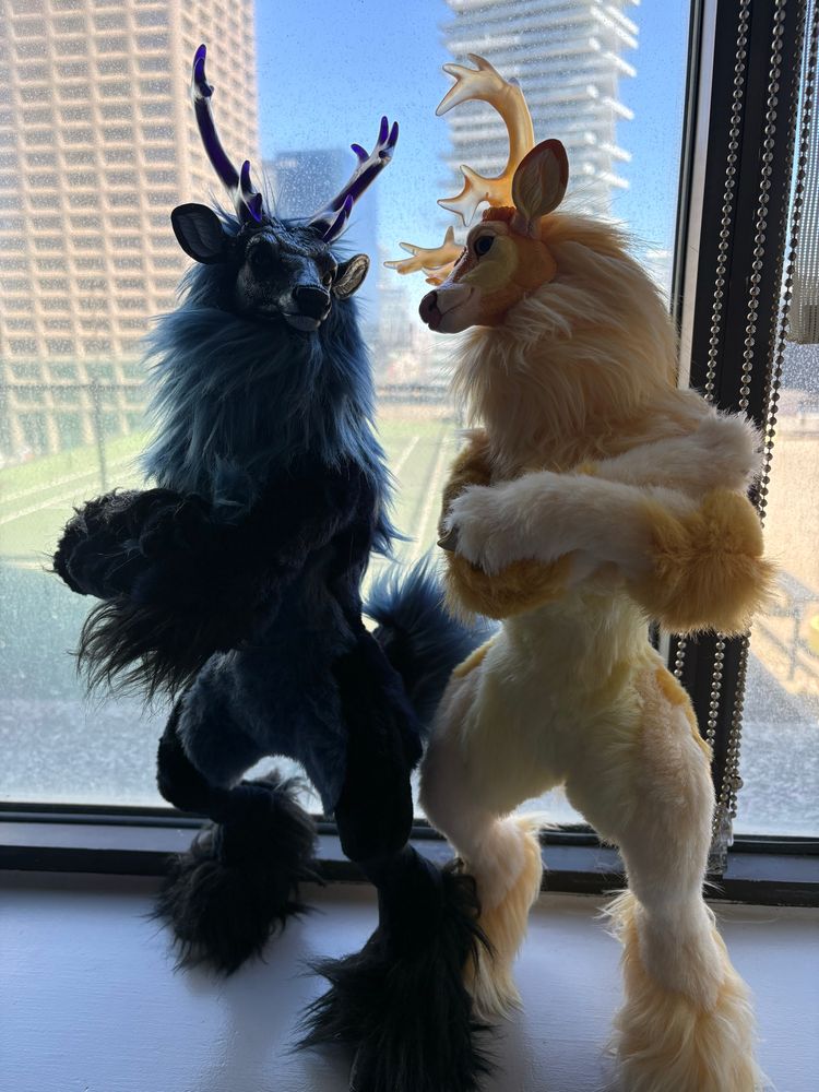 The same two art doll deer posing in a window. Their arms are crossed and they are looking at each other. The blue is on the left further back while the yellow one is closer to the viewer.  