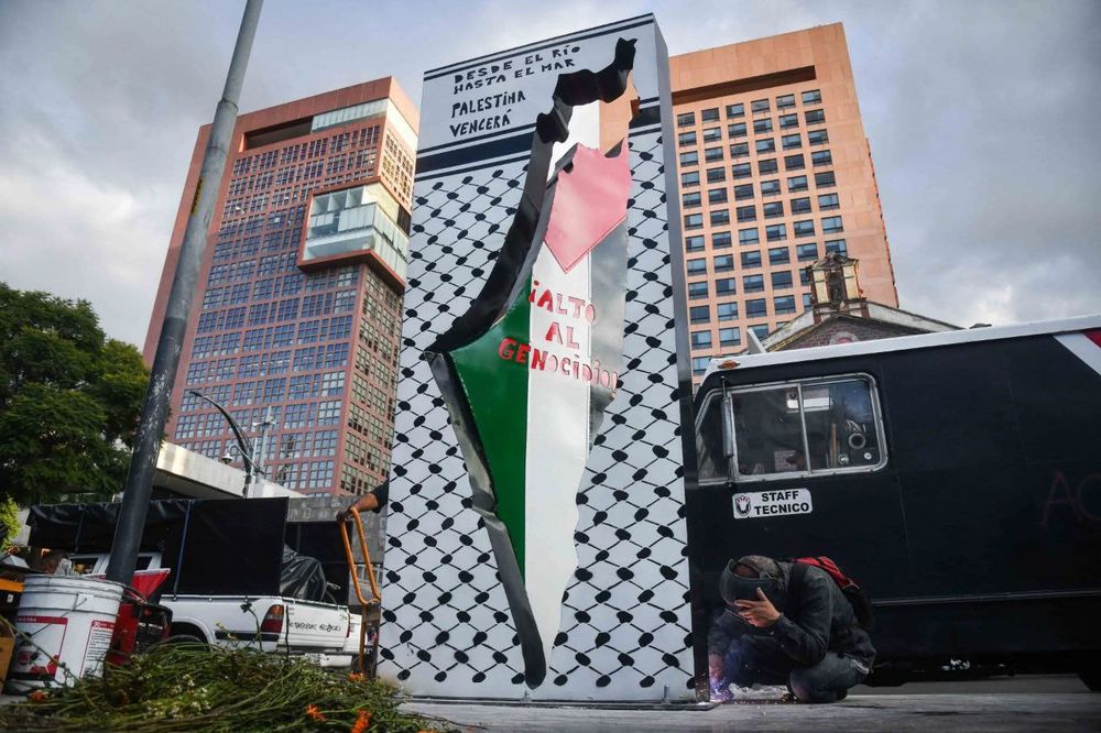 A bottom-up photo of the new anti-monument to Palestine in Mexico City, a rectangle shaped piece with text at the top that reads "Desde el río hasta el mar, Palestina vencerá" with white and black patterns resembling a keffiyeh and in the middle the map of Palestine opens towards the back, as if a door, painted in the colors of the Palestinian flag, on which reads "Alto al genocidio!"