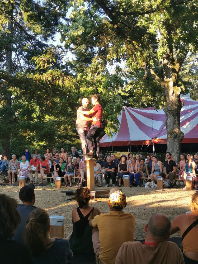 Un duo de circassiens en équilibre sur une planche, au milieu du public assis.