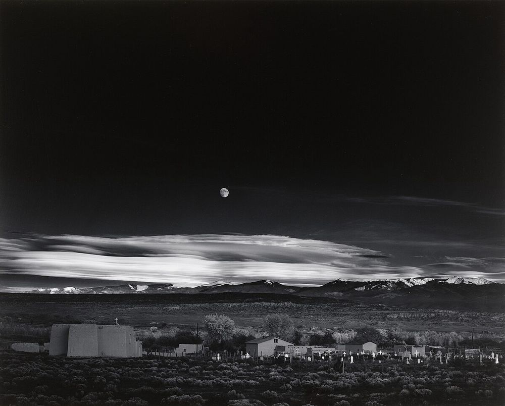 Moonrise over Hernandez - A black and white photo of a desert landscape in the American Southwest in the early evening with the moon rising in a darkened sky over a few houses in the distance.