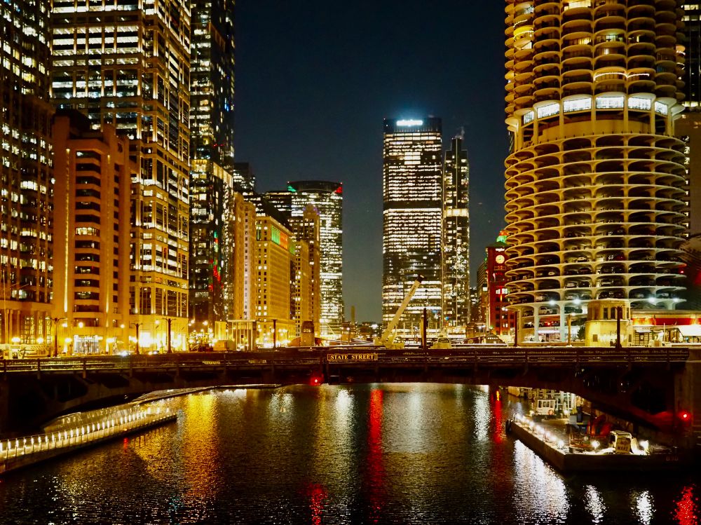 View of tall buildings along the Chicago River at night in winter.  The buildings are lit with warm yellow lights which reflect in the water of the river.