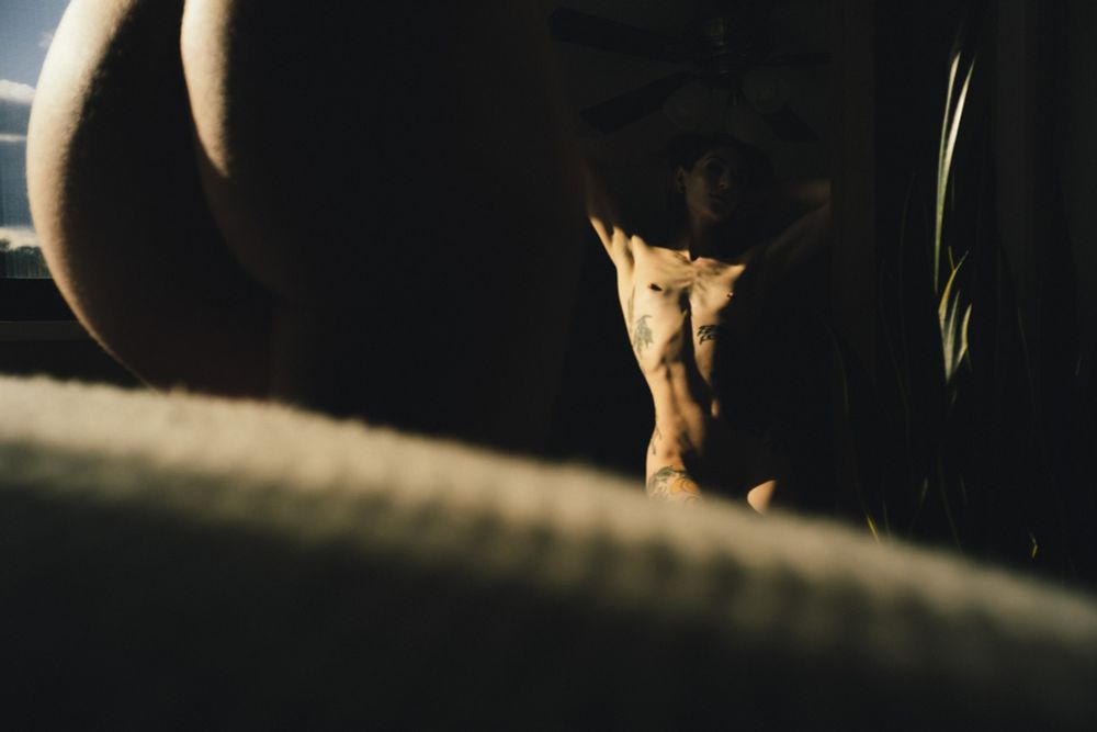 Artistic nude portrait of a tattooed woman standing in front of a mirror, with golden sunlight casting dramatic highlights across her torso. The foreground shows a close-up of her hip and thigh, blurred and silhouetted against the light, creating a layered composition. Her reflection in the mirror reveals her full pose, arms raised behind her head, surrounded by shadows, a ceiling fan, and a tall houseplant — blending introspection with bold visual contrast.

