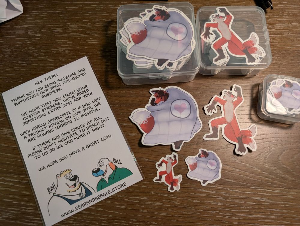 Photo of many sticker prints using the custom artwork from this post (orange fox curled up under weighted blanket and fox sprawled out on back blushing and tail wagging).

To the left lays a thank-you note from Bear and Beagle store for having ordered, offering support, etc.