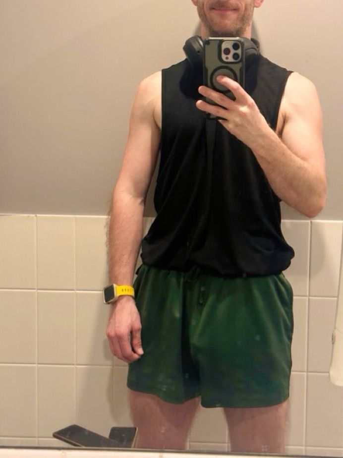 A young male is standing in a toilet change room. He is wearing a black muscle tank and green shorts, which due to free balling, the outline of the head of his cock is visible through his shorts 