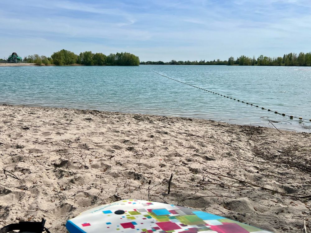 Man sieht einen Teil eines Bodyboards am Rande eines sonnenbeschienenen Sees.