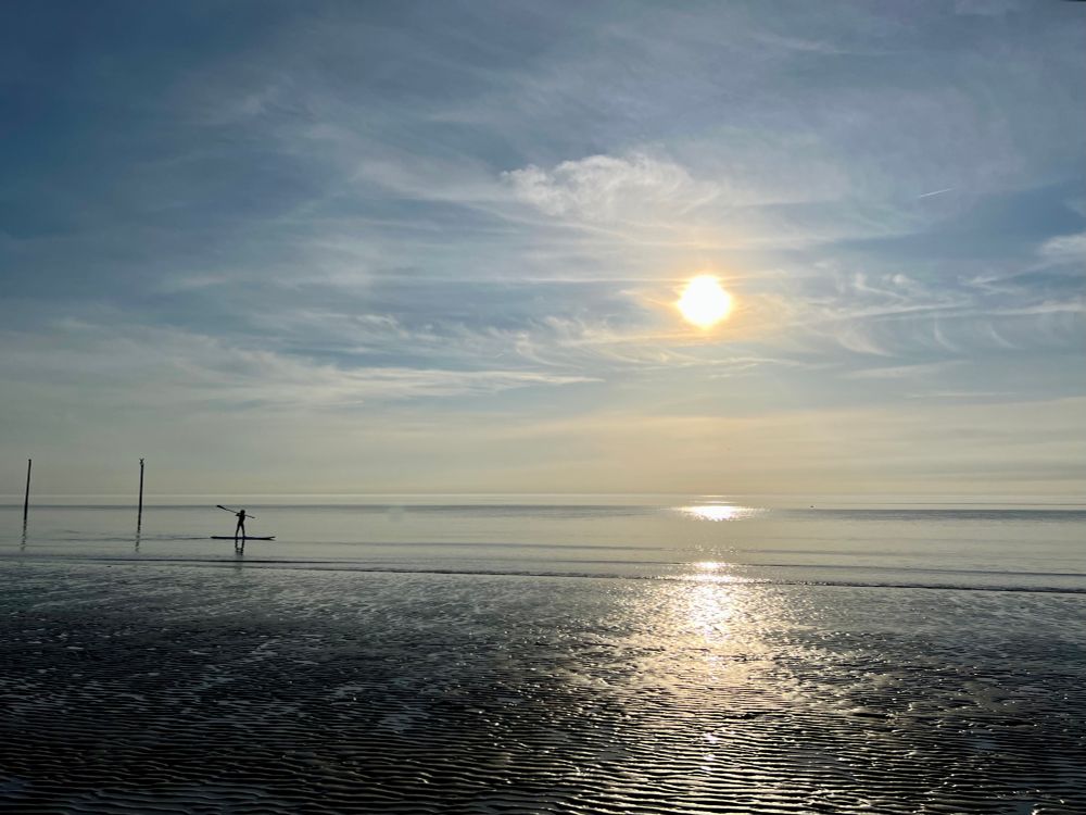 Zu sehen ist das Meer, genauer die Nordsee. Die Sonne steht schon relativ tief. Es gibt starke Reflektionen auf dem Wasser. Im Vordergrund ist etwas vom Wasser geformter Strand zu sehen, es scheint gerade Niedrigwasser zu sein. Links am Bildrand fährt ein Kind auf einem Stand Up Paddelboot.
