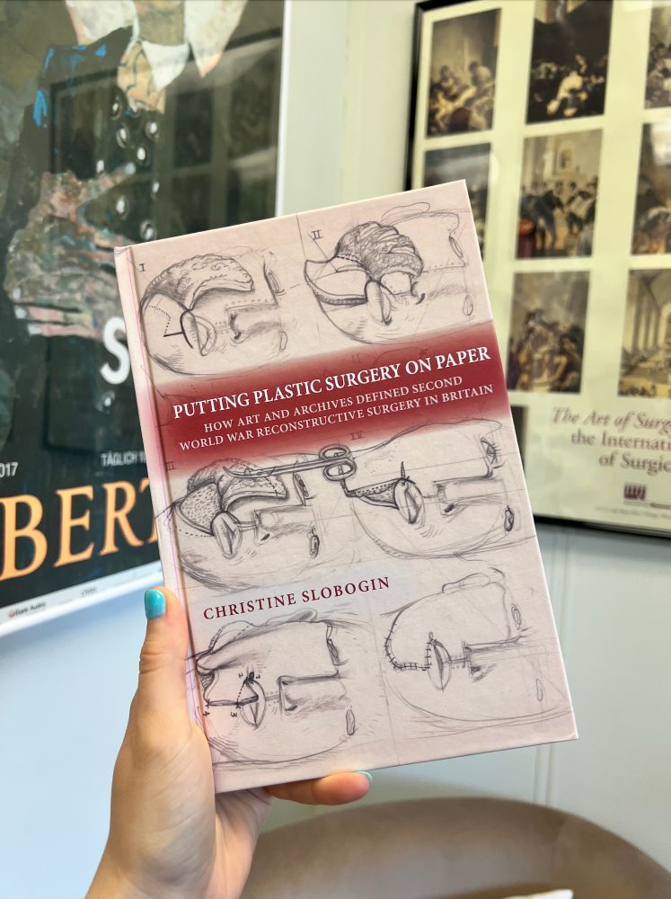 Holding the hardcover version of _Putting Plastic Surgery on Paper_ up in front of two framed posters. The cover of the book shows a face being surgically reconstructed, in six steps, drawn with pencil on beige paper. The title (in caps) is backed by a dark red (the full title is Putting Plastic Surgery on Paper: How Art and Archives Defined Second World War Reconstructive Surgery in Britain).