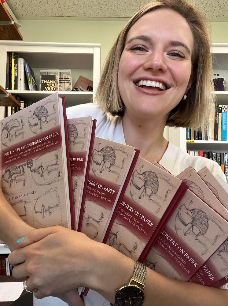 Happy author (me) holding a bunch of hardcover copies of _Putting Plastic Surgery on Paper_. I am a white blonde woman with short hair wearing a white top and in an office with bookshelves visible in the background.