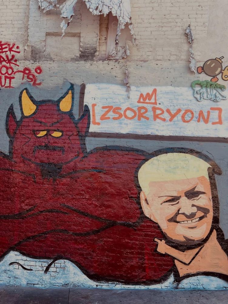 Satirical mural in Los Angeles, USA, showing Donald Trump smiling while lying in bed with a devil-like figure. The devil has red skin, yellow horns, and a muscular build. Above the scene is the graffiti tag “[ZSORRYON]” with a crown symbol. The piece references the July 2025 South Park episode where Trump is literally portrayed in bed with the devil, critiquing his political persona and alliances through animated satire.