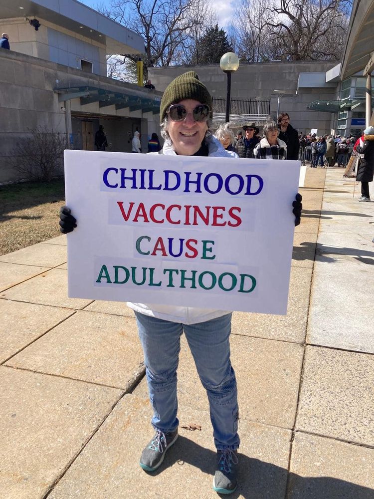 A smiling person wearing sunglasses, a green beanie, and casual clothing holds a humorous protest sign outdoors that reads, 'CHILDHOOD VACCINES CAUSE ADULTHOOD,' parodying typical anti-vaccine slogans. The scene takes place in a sunny area with several people visible in the background, suggesting a gathering or rally.