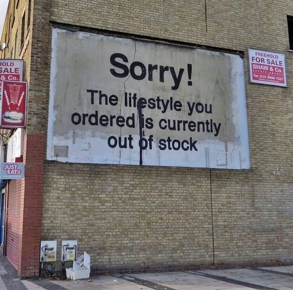 A satirical mural attributed to Banksy on the side of a brick building, featuring bold black text on a white background that reads, "Sorry! The lifestyle you ordered is currently out of stock." The piece is situated on a weathered wall, surrounded by signage for real estate and a nearby fast-food delivery service, emphasizing themes of consumerism and modern societal disillusionment.