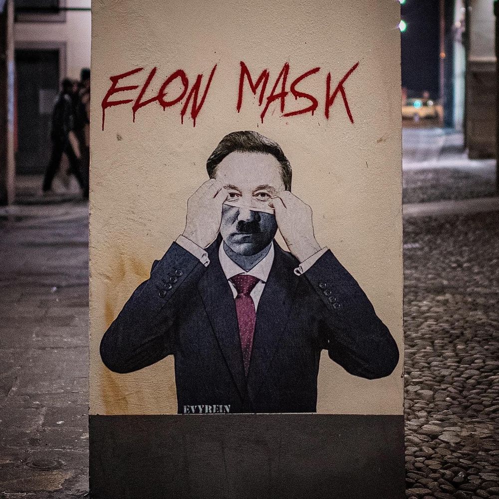 Street art titled "Elon Mask" by Evyrein, condemning Elon Musk’s Nazi-style salute during Trump’s 2025 inauguration and his later appearance at a German far-right AfD event. The artwork shows Musk pulling a black mask over his face, mimicking Hitler’s mustache, with bold red graffiti text "Elon MASK.